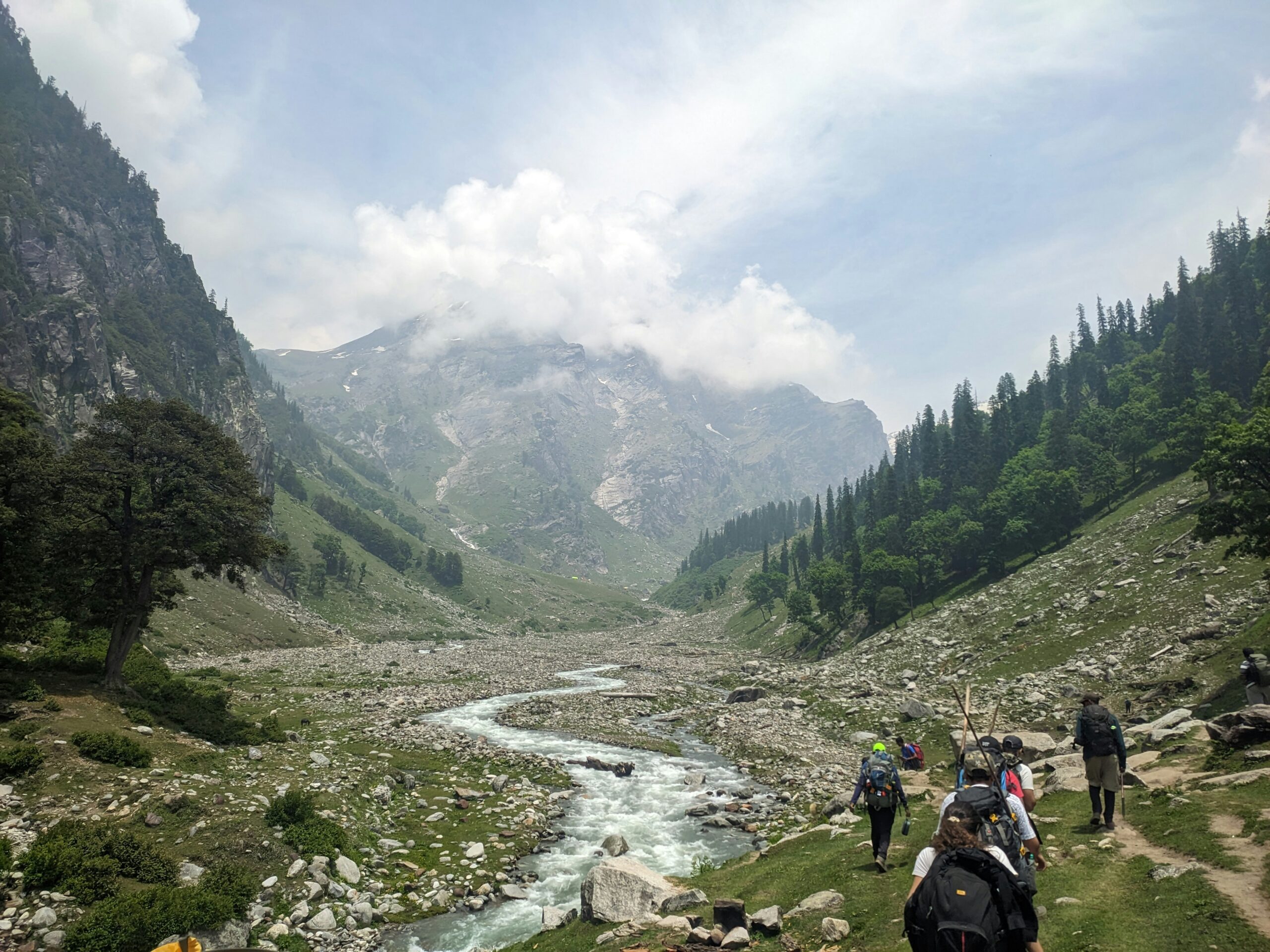 Group of trekkers walking beside a mountain stream in a green valley during Hampta Pass Trek in Himachal Pradesh